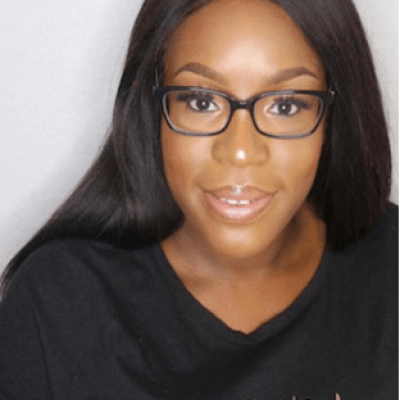 A head and shoulders shot of Helen Ayo-Ajayi wearing glasses and a black t shirt stood in front of a white wall looking directly into the camera
