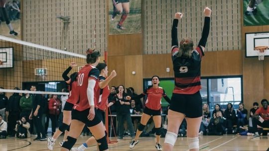 Five women in Volleyball clothing jumping around in excitement and celebration. The women are in a sports hall with spectators all around them.