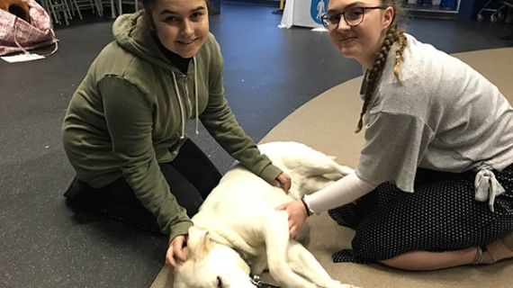Two students smile at the camera whilst they stroke a guide dog who is laid on the floor